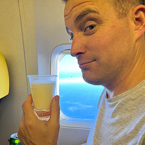 A smirking male poses with a drink on an airplane