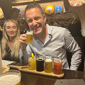  A smiling man poses with five multicoloured beers next to a female