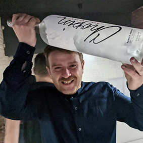 A man smiles while holding a large bottle of vodka inside a bar