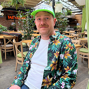  A man wearing a floral shirt and a baseball cap sits outside a bar