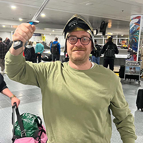  A man wearing spectacles and a knights helmet holds a sword in an airport