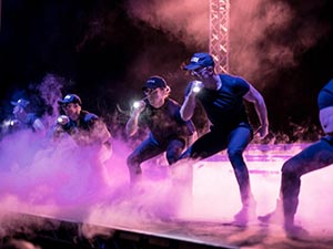 A line of four men dancing on stage and holding torches up, to a smoky backdrop