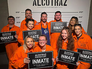 A group of people wearing orange jumpsuits and holding inmate signs, standing in front of a prison style height wall. 