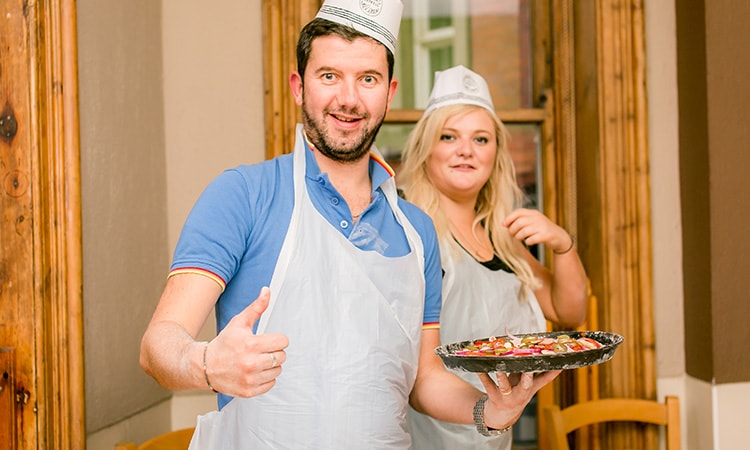 A man in a chef hat and apron, holding up a pizza with a woman in the background