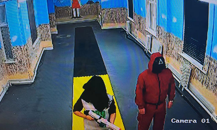 A security camera image of a blindfolded person holding a Nerf gun with a guard next to them