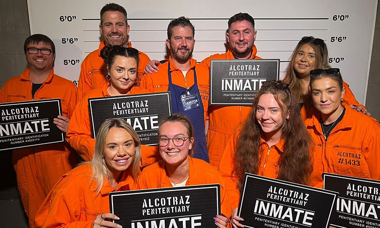 A group of people wearing orange jumpsuits and holding inmate signs, standing in front of a prison style height wall. 