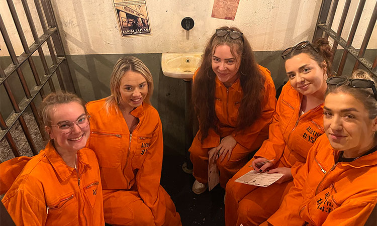 A group of women wearing orange jumpsuits sitting in a cell smile at the camera in Alcotraz in Bristol. 