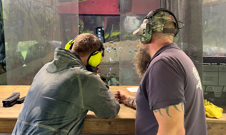 Two men wearing ear defenders and looking towards a shooting range as one of them aims a gun. 