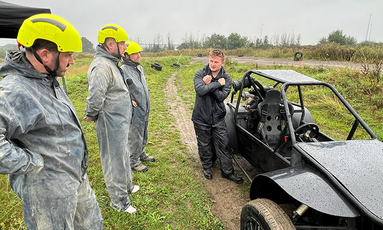 Three men in jumpsuits and helmets listening to an instructor in front of rage buggies. 