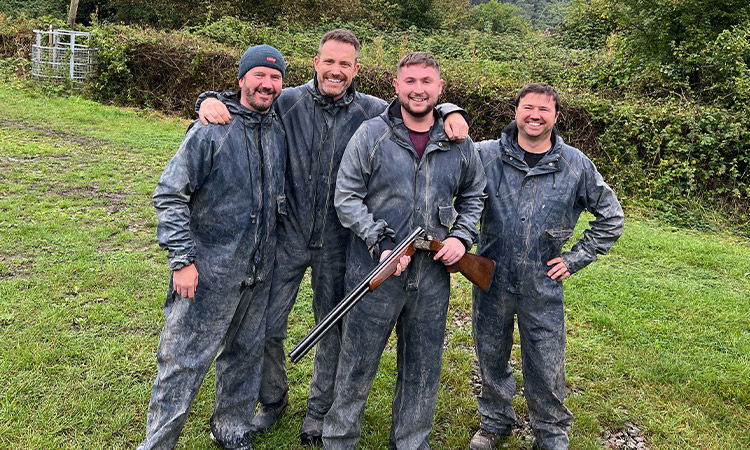 Four men posing for a camera wearing jumpsuits while one holds a shotgun. 