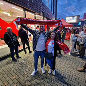  Two people posing for the camera in front of a football stadium with red and white scarves
