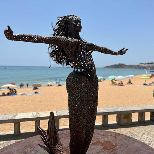 A metal statue of a mermaid with the beach in the background