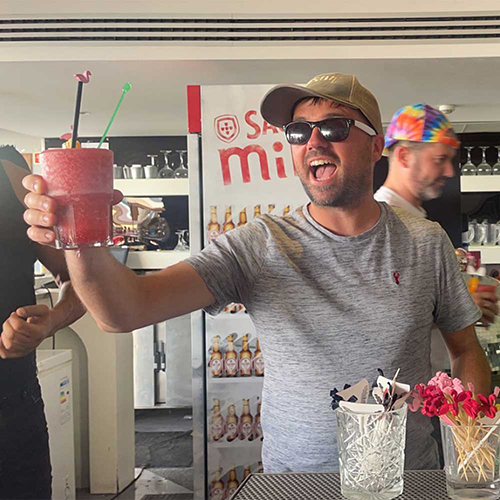 A male member of our team wearing sunglasses and holding up a red cocktail he has just made 