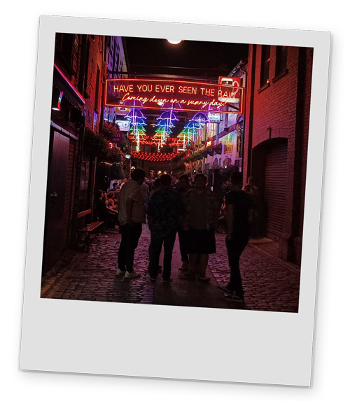An alley at night-time, with a neon sign that says Have you ever seen rain coming down on a sunny day and colourful umbrella decoration in the background