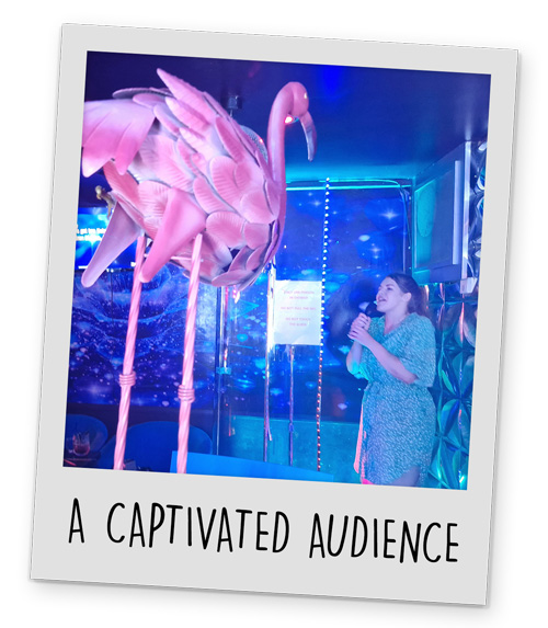 A giant fake flamingo looking down on a female member of our staff singing karaoke in a karaoke booth