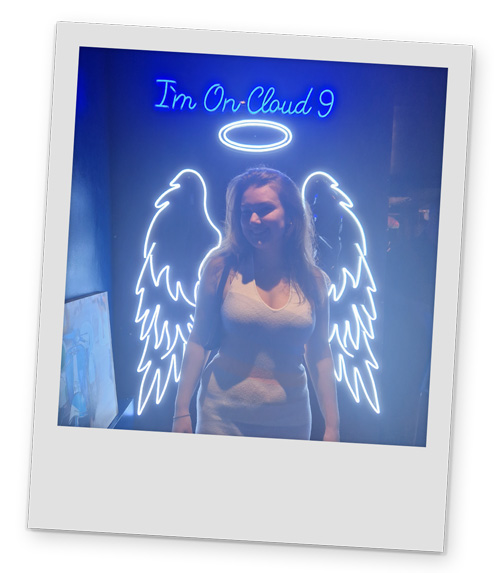 A female member of staff posing in front of a sign that says 'I'm on Cloud 9. She has an angel outline in lights.