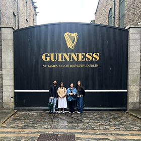 Four members of staff posing in front a massive black gate that reads Guinness, St James's Gate Brewery Dublin 