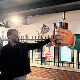  Akash doing a fist bump with a cartoon statue of a man who is holding a Guinness, in the advertising room of the Guinness Factory Tour 