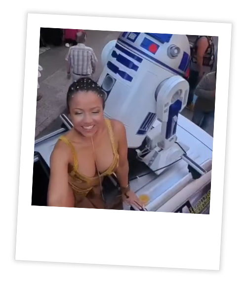 A Polaroid of a woman with a low cut top who is stood next to R2-D2