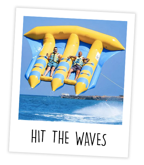 A Polaroid of two men on a flying fish which is a flat-shaped inflatable that is being pulled behind a speed boat and flying in the air