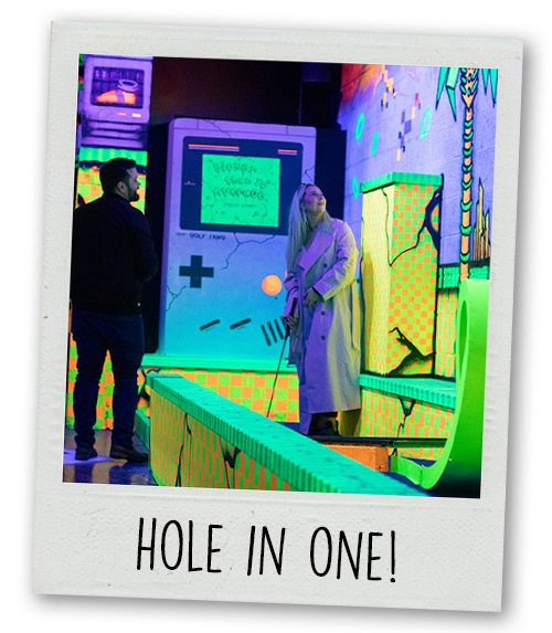 A Polaroid of a man and a woman playing a gaming-themed mini golf hole with the caption Hole In One!