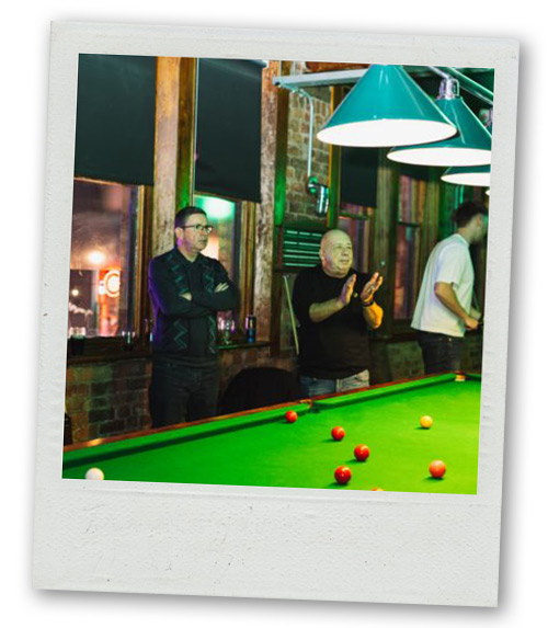 A Polaroid of two men stood beside a pool table with one of them clapping and the other with his arms folded