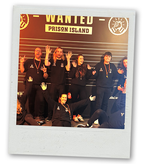 A Polaroid of a mixed gender group posing in front of a backdrop that reads Wanted Prison Island