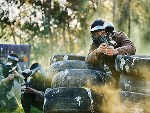 A man paintballing who is hiding behind a stack of tires and aiming his paintball gun 