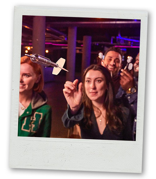 A Polaroid of a woman throwing a dart at a dartboard whilst her friends watch on