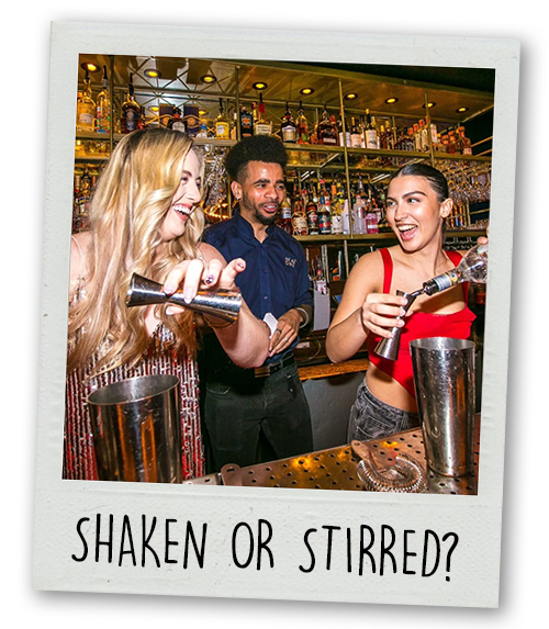 A Polaroid of a male bartender teaching two women on a hen do how to make a cocktail with the caption Shaken Or Stirred?