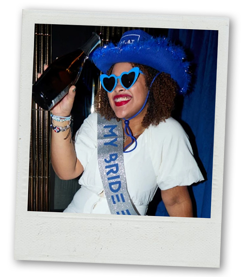 A Polaroid of a woman wearing a cowboy hat and a hen do sash who is holding up a bottle of drink