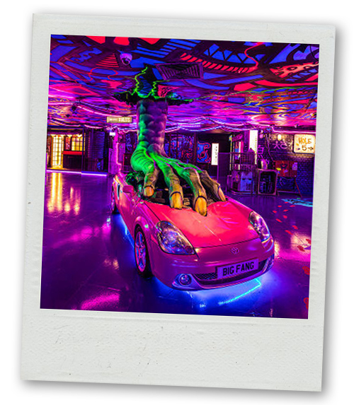 A Polaroid of a monstrous looking foot coming out of the ceiling and crushing a pink car which has the number plate Big Fang
