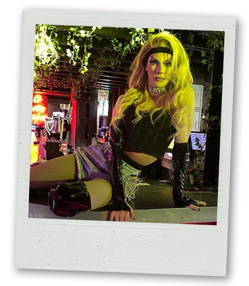 A Polaroid of a drag queen in a long blonde wig sitting on top of a bar