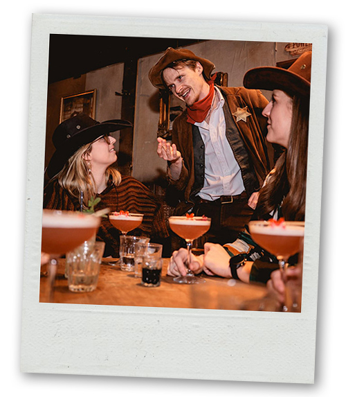 A Polaroid of two women wearing cowboy hats interacting with an actor dressed up as a cowboy whilst they drink cocktails