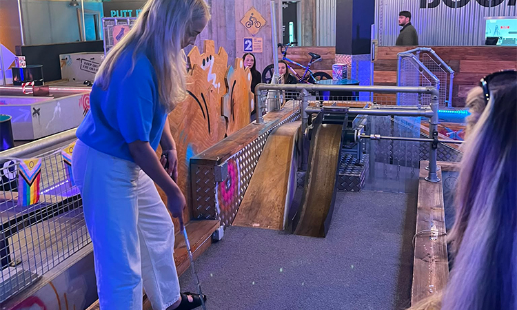 A blonde woman about to hit a golf ball up a ramp as a part of crazier golf