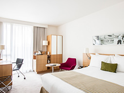 A double bed made up in a hotel room, with wardrobes, a desk and chairs in the background