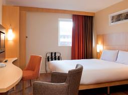 A white double bed in a hotel room, featuring a desk, TV, floor length red curtains and two chairs