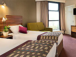 Two double beds in a hotel room, topped with throws and cushions, with a green sofa in the background