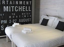 A double bed topped with black cushions and towels, with a black and white wall in the background