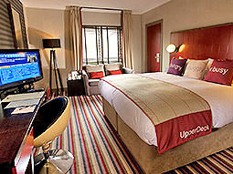 A double bed in a hotel room, topped with a throw and three cushions, facing a turned on TV, desk and desk chair with a sofa in the back