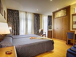 A large double room with blue bedding and curtains, with dark wood furniture and paintings on the wall 