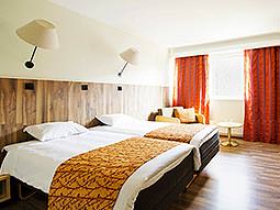 A spacious twin guest room with wall lamps and red curtains