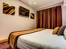 A double bed with a throw and cushions on top, facing a flat screen TV on the wall