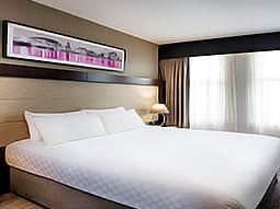 A large double bed with white bedding, in a room with a large black, white and pink landscape canvas on the wall