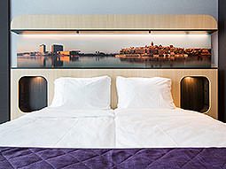 A white double bed, topped with a purple throw, with a pitcure above the bed