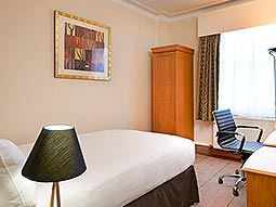 A lamp in the foreground of a double room with a desk chair and a small, thin wardrobe 