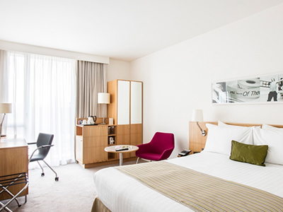 A double bed made up in a hotel room, with wardrobes, a desk and chairs in the background