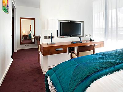 A double bed with a blue throw, facing a desk and TV with a mirror on the wall in the back