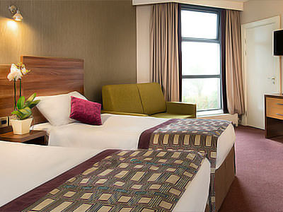Two double beds in a hotel room, topped with throws and cushions, with a green sofa in the background and drawers