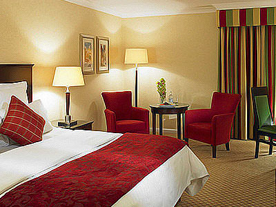 Double bed in a hotel room, topped with a red throw and cushion, with two red chairs, a table and desk in the background